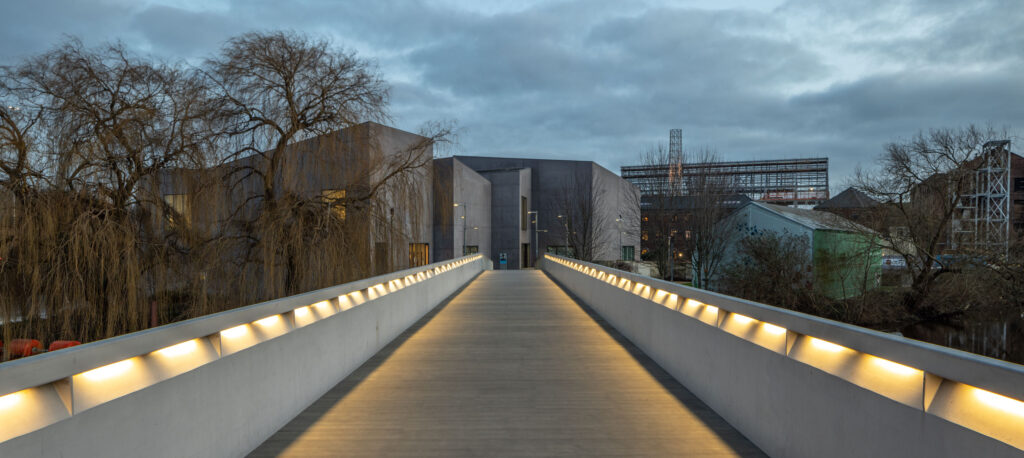 Illuminated concrete bridge leading to modern architectural building, flanked by trees and industrial structures under cloudy sky.