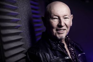 Bald man with a goatee and earring, wearing a leather jacket, smiles in front of acoustic foam panels with blue lighting.
