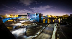 Night view of a flowing river with a modern blue-lit pedestrian bridge and buildings illuminated in the background.