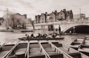People enjoying a punt ride on a river beside a bridge, with historic buildings and parked boats in the background.