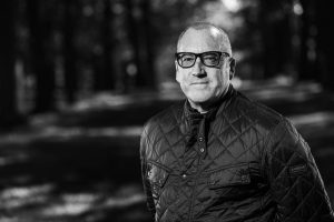 Man with short hair and glasses standing in a park, wearing a quilted jacket, black and white photo, trees blurred in the background.