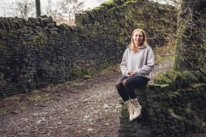 Woman in gray hoodie sits on moss-covered stone wall, smiling in a rustic, outdoor setting with an old stone pathway.