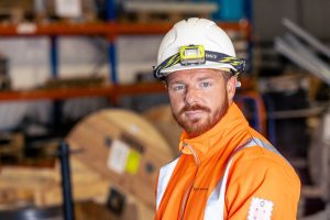 A construction worker in an orange high-visibility jacket and helmet with a headlamp stands in a warehouse, looking at the camera.
