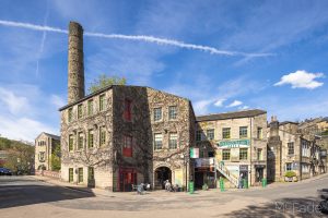 Historic stone building with a tall chimney, green windows, and a cafe at the base under a clear blue sky in Hebden Bridge.