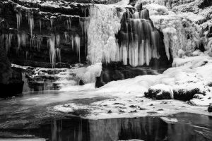 Frozen waterfall with icicles hanging from snowy rock formations, reflecting on the icy water below. Monochrome image.