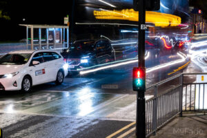 Long exposure shot of Leeds cityscape at night with light trails from passing vehicles on a wet road.