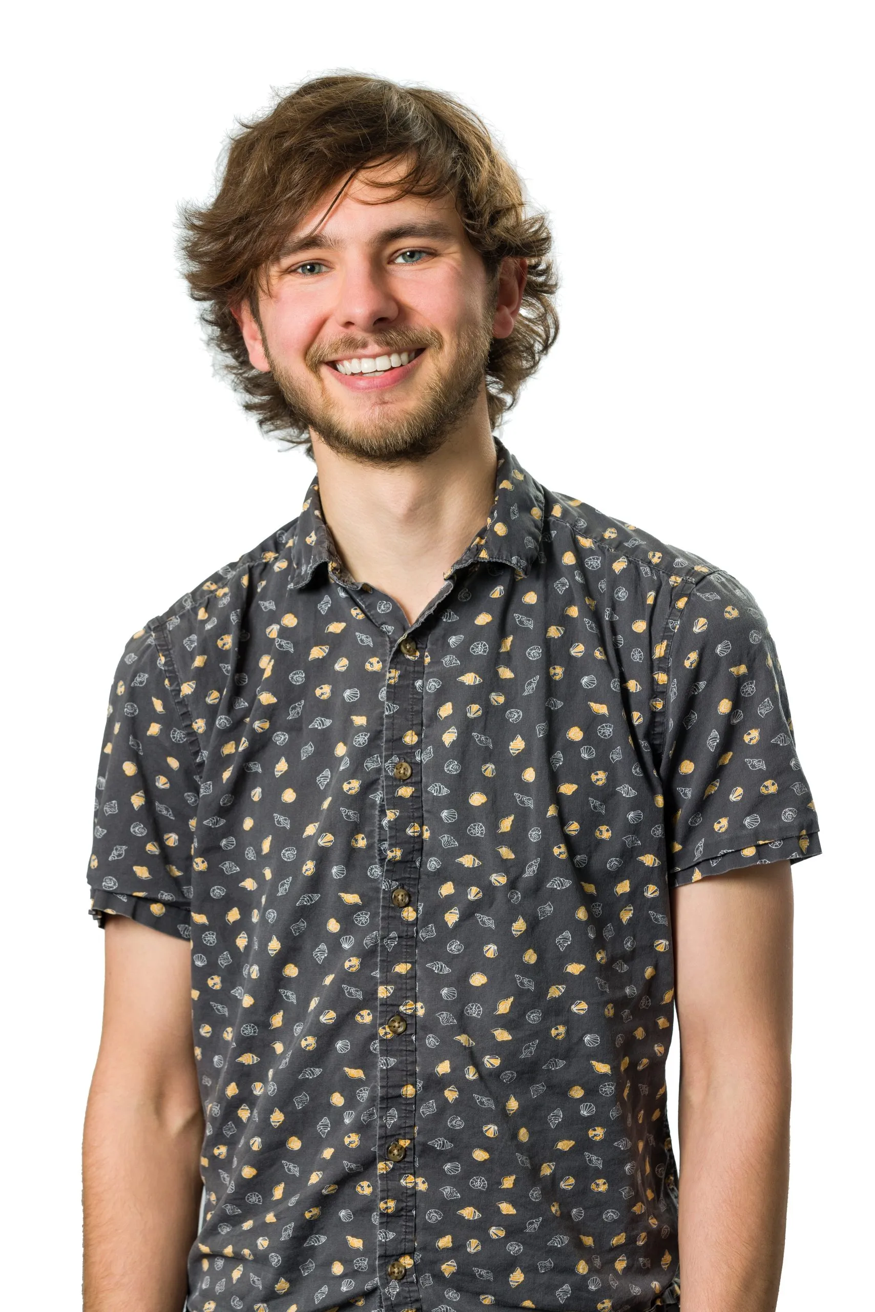 Smiling person with wavy brown hair, wearing a dark patterned shirt, against a white background.