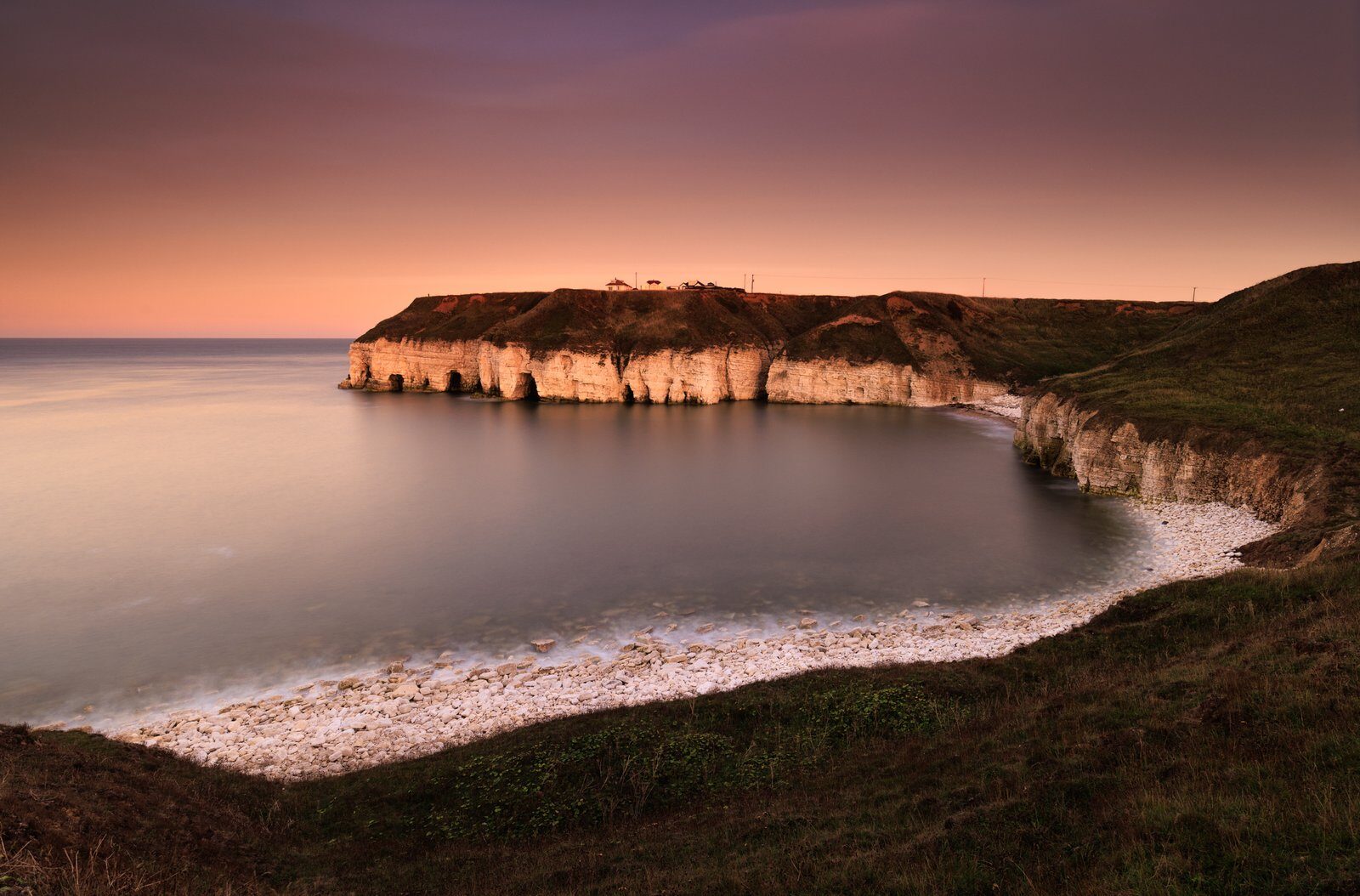 A curving chalk-cliff bay with a pale pebble beach, smooth sea and orange-pink evening sky.