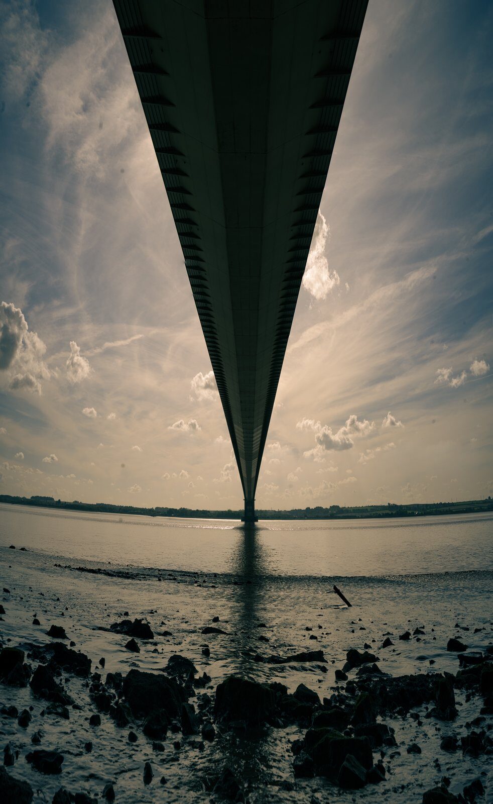 A fisheye photograph looking along the underside of the Humber Bridge, with shoreline and sky.
