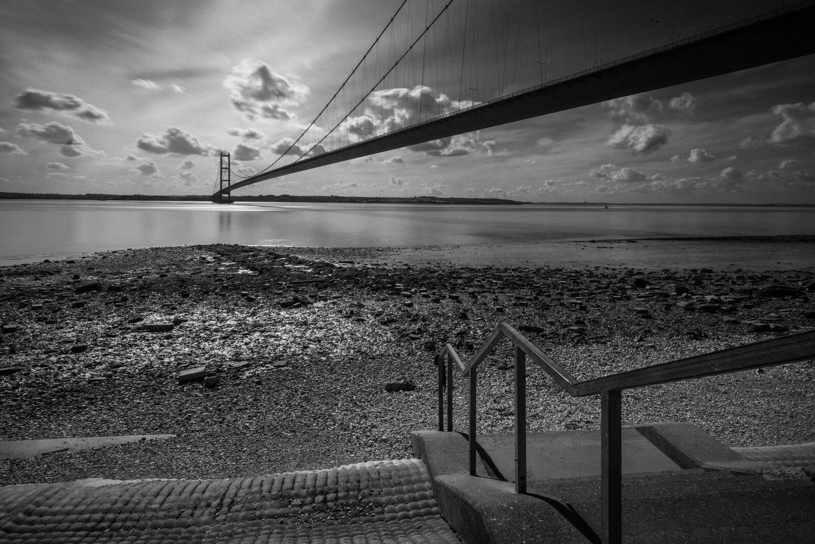 A black-and-white photograph of the Humber Bridge sweeping over the river, with steps and a handrail in the foreground.