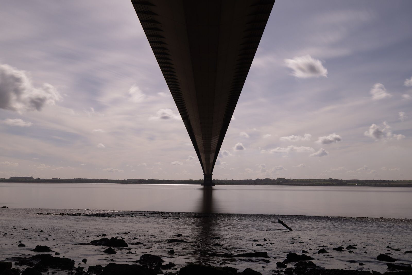 The Humber Bridge deck narrowing towards its support above smooth water and a rocky shoreline.