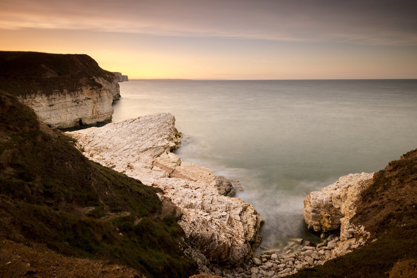 White chalk cliffs and foreground rock above smooth water, beneath a warm evening sky.