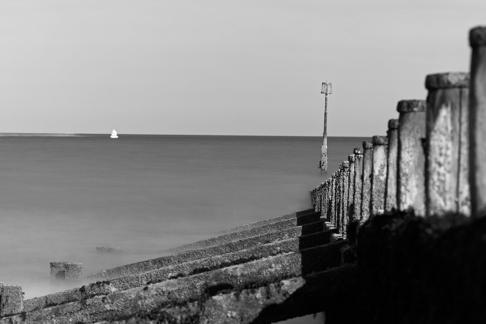 A black-and-white view along a weathered beach groyne, with smooth water, a tall marker and a small distant boat.