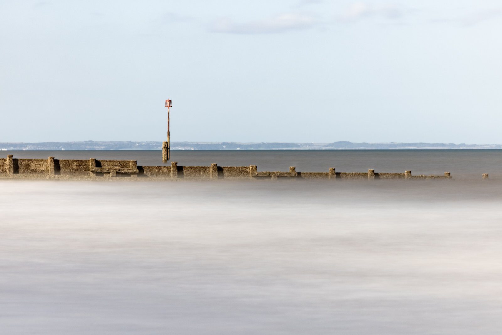 A beach groyne stretching across smooth pale water, with a tall marker rising against a blue sky.
